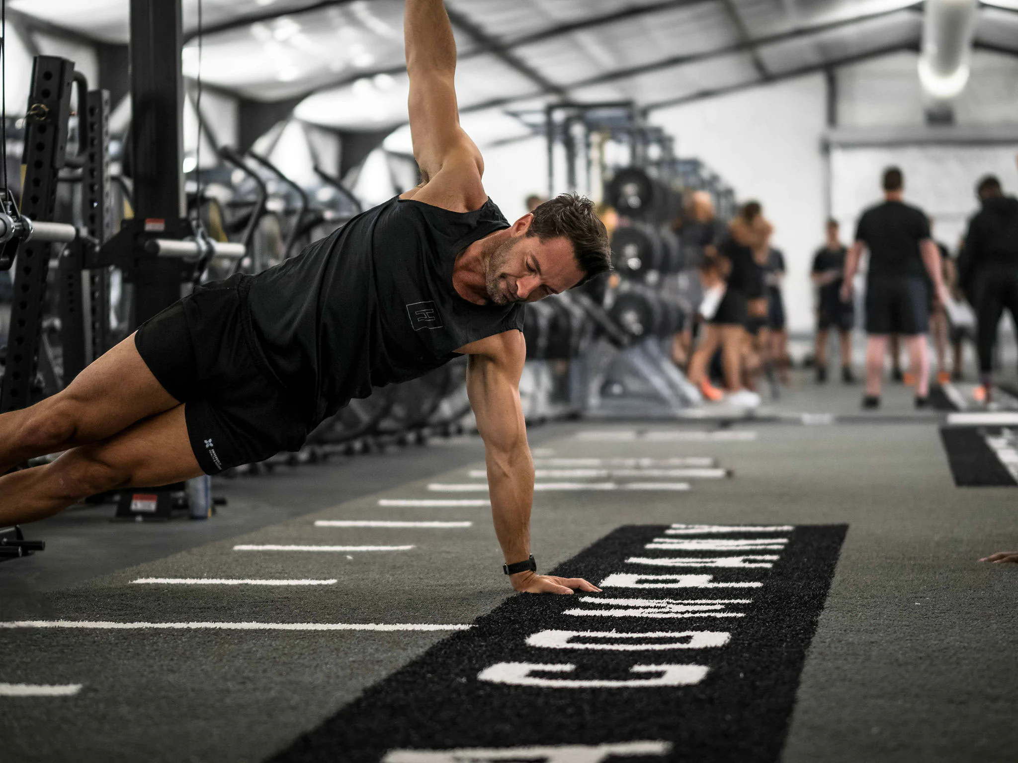 A man in athletic wear performs a one-arm side plank on a gym floor, surrounded by weights and exercise equipment, with other people working out in the background.