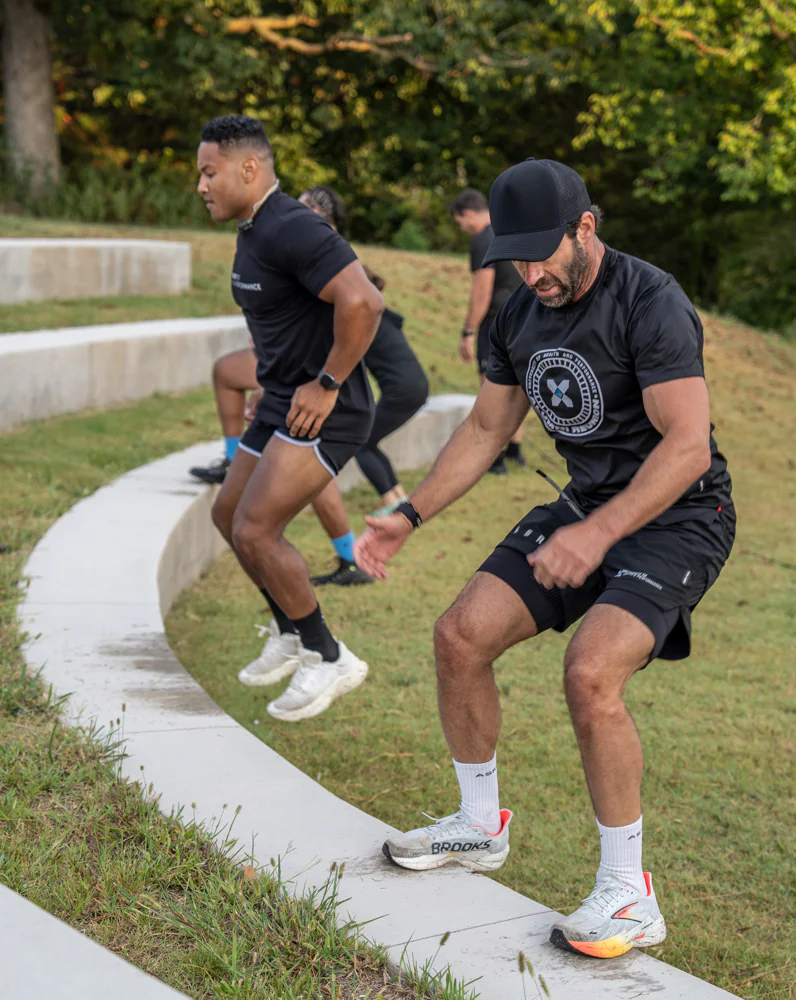 Two men in athletic wear exercise outdoors, stepping or jumping along curved concrete steps in a grassy area, with more people and trees in the background.