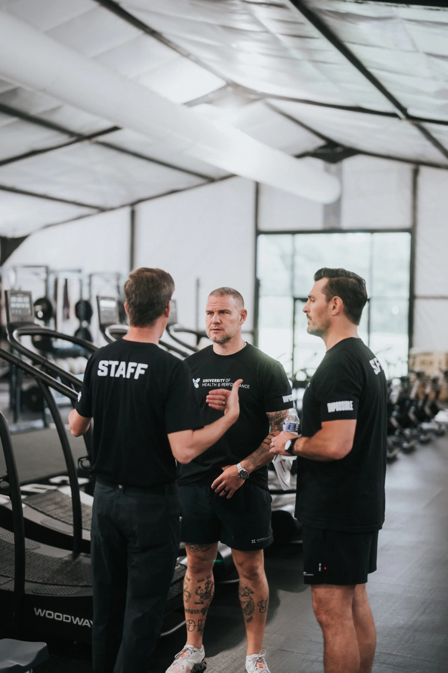 Three men in black athletic wear, with STAFF written on their shirts, stand together talking in a gym with treadmills and workout equipment in the background.