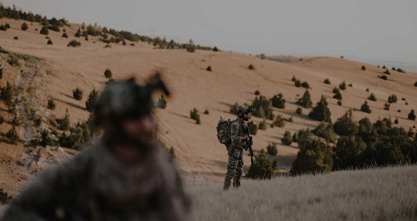 Two soldiers in camouflage gear stand in a grassy, hilly landscape with sparse trees. One is in focus in the distance while the other is blurred in the foreground. Both appear alert and equipped for a mission.
