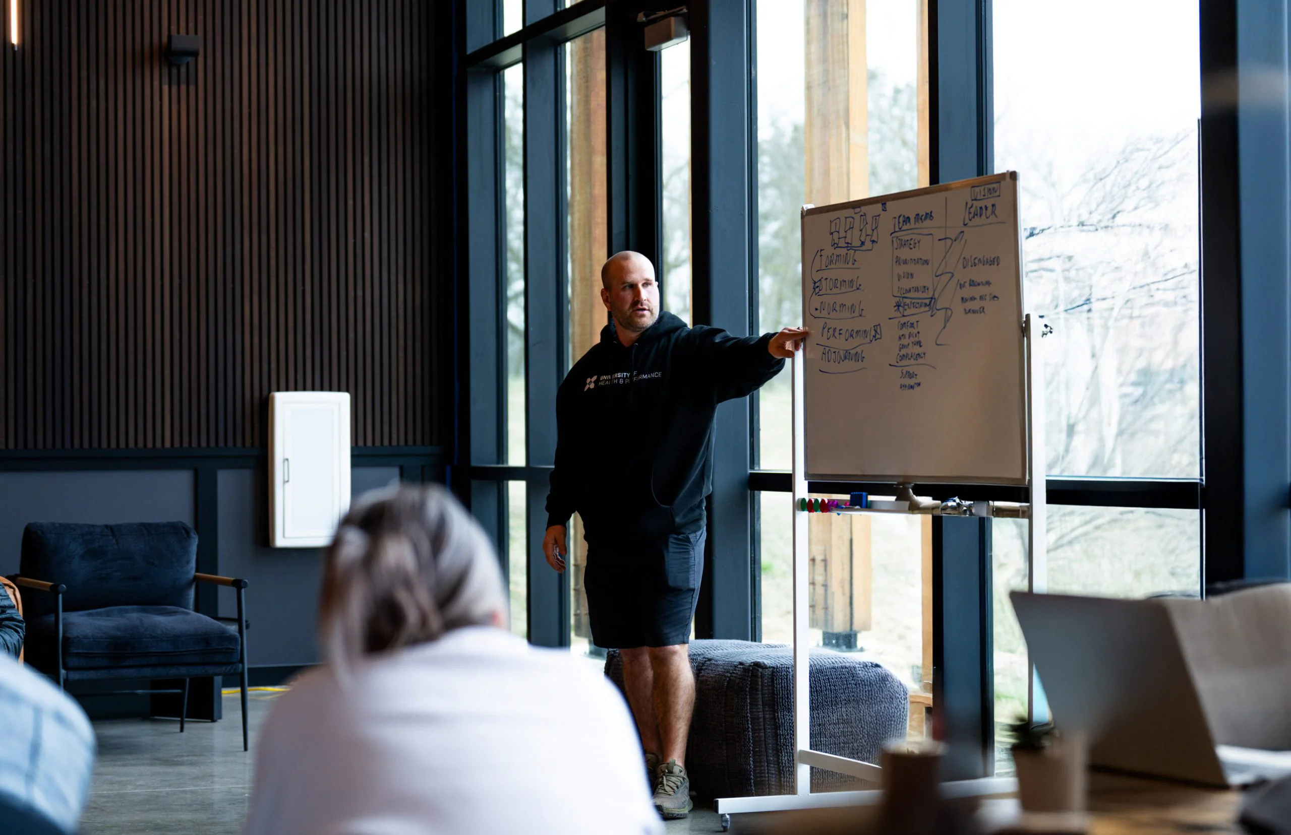 A man in casual clothes stands by a whiteboard, pointing at notes, while a seated person listens in a modern room with large windows and natural light.
