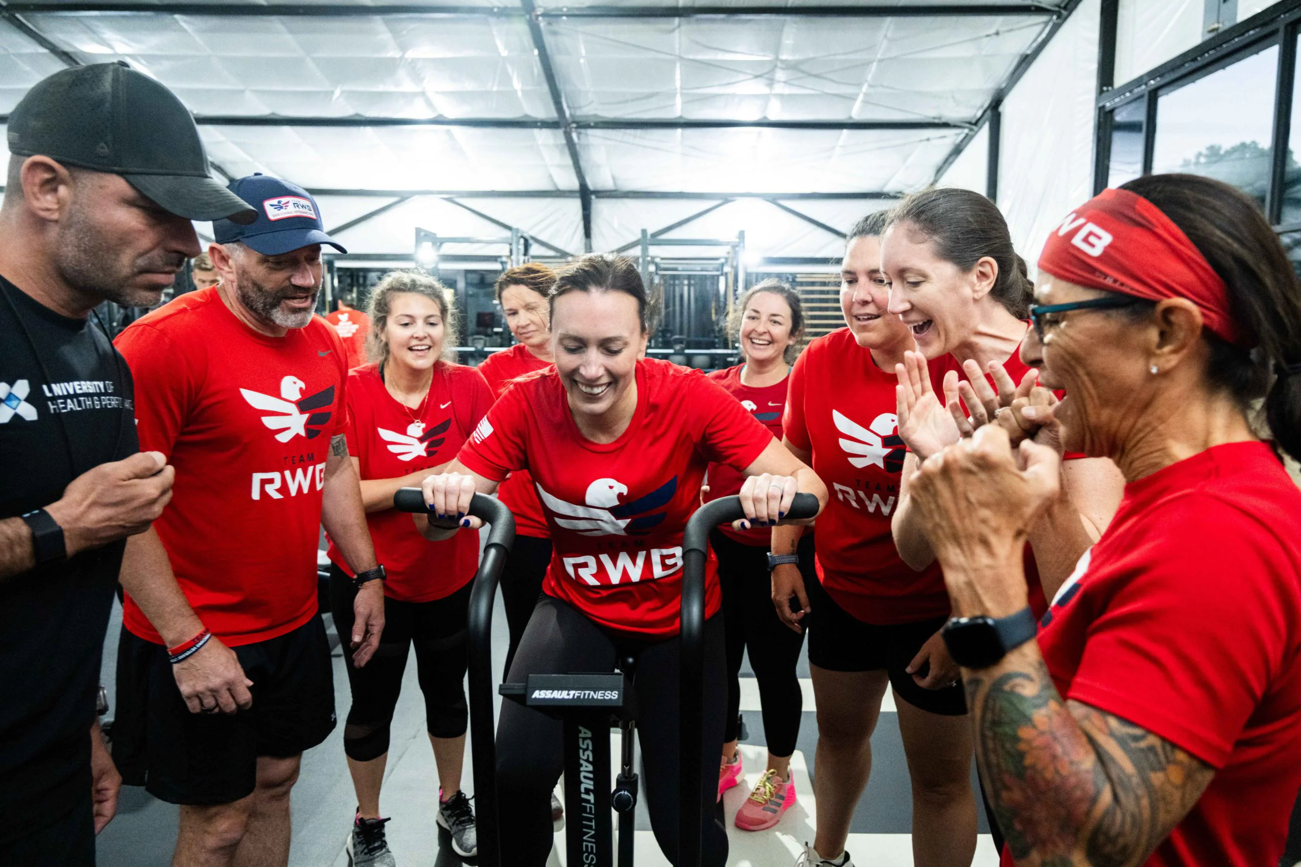 A group of people in matching red shirts cheer and support a woman exercising on a stationary bike inside a gym, creating a lively and encouraging atmosphere.